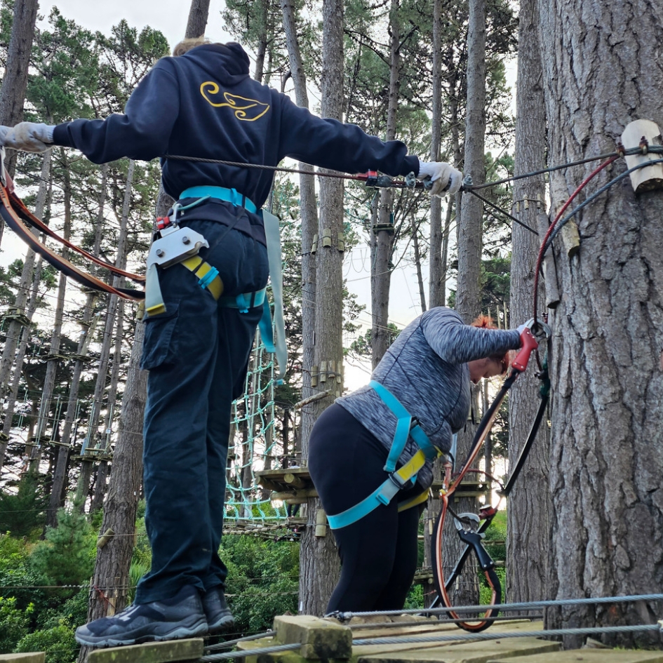 A boy in a climbing harness watches Kitty Fitton climb up to a platform that is in the trees. behind them is a forest with a rope course.