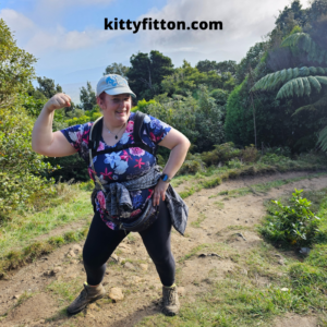 Shows kitty fitton in walking gear posing on a track with distant sea and hills in the view behind her. She is tired but smiling. 